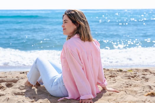 portrait of a happy young woman sitting on a sandy beach, smiling at the camera with the ocean behind her. Relaxed vibe, casual outfit, natural light and peaceful outdoor setting.
