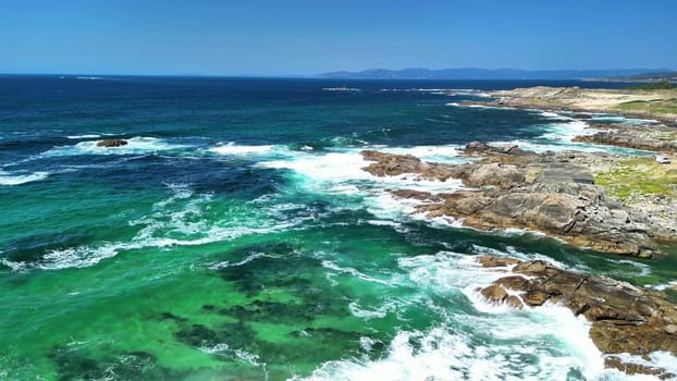 An aerial shot beautifully captures the waves crashing against the rugged rocks along a vibrant coastline