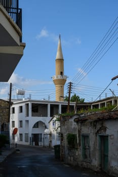 A quiet street in the historic town of Lefke, Cyprus, with white traditional buildings and a tall mosque minaret visible in the background.