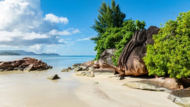 A peaceful beach with soft sand, large rocks, and vibrant green trees against a calm blue ocean. Praslin, Seychelles.