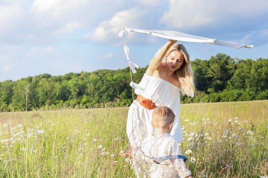 In a meadow in the grass and wildflowers, a child and a mother in a white dress and with blond hair are launching a kite into the sky, having fun with their little son. High quality photo