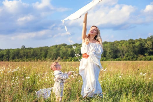 In a meadow in the grass and wildflowers, a child and a mother in a white dress and with blond hair are launching a kite into the sky, having fun with their little son. High quality photo
