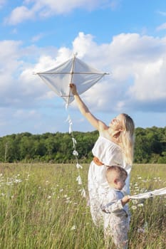 In a meadow in the grass and wildflowers, a child and a mother in a white dress and with blond hair are launching a kite into the sky, having fun with their little son. High quality photo