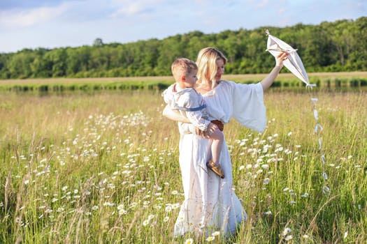 In a meadow in the grass and wildflowers, a child and a mother in a white dress and with blond hair are launching a kite into the sky, having fun with their little son. High quality photo