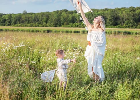 In a meadow in the grass and wildflowers, a child and a mother in a white dress and with blond hair are launching a kite into the sky, having fun with their little son. High quality photo