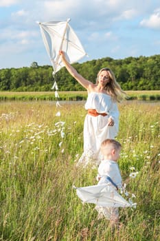 In a meadow in the grass and wildflowers, a child and a mother in a white dress and with blond hair are launching a kite into the sky, having fun with their little son. High quality photo