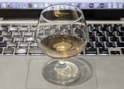 Brandy snifter or Whiskey glass stands on Laptop with laptop screen in the background. use it as your Wallpaper, Poster and Space for text, Selective focus.