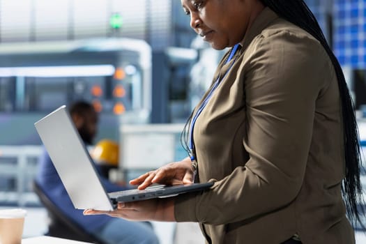 Black alternative energy consultant observes operations in a solar tech plant, ensuring factory practices meet green energy targets. Specialist uses laptop for manufacturing efficiency.