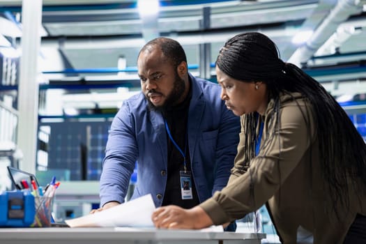 Black team of engineers technicians share ideas in a solar panel factory, reviewing blueprints and sharing ideas to enhance renewable energy production and sustainable innovation.