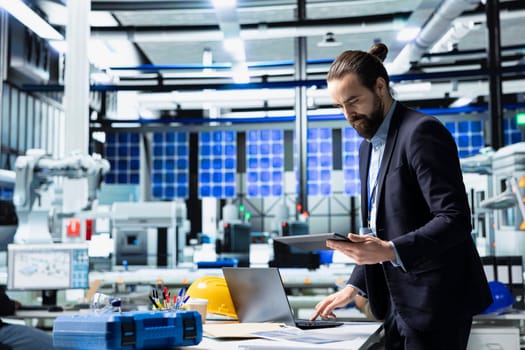 Solar energy specialist in a facility reviews data on gadget, inspecting manufacturing plant performance and symbolizing solar innovation. Industrial employee oversees production line.