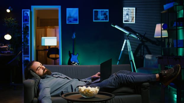 Exhausted man sleeping on couch after working on laptop all day before managing to watch TV and eat popcorn. Freelancer falls asleep while solving tasks on notebook and watching film, camera B