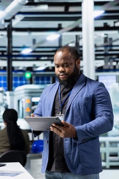 African american business person walks through modern factory inspecting solar panel machinery, reading important insight for the corporate commitment to sustainability and renewable power.
