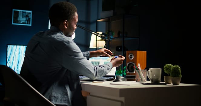 Black man organizing his schedule with a planner and putting a reminder, creating an adhesive note post it and browsing the web. Modern productive guy writes a memo for research. Camera A.