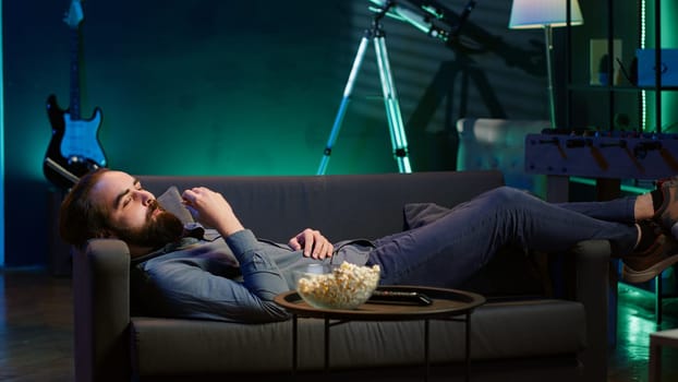 Smiling man lounged on couch at home eating popcorn while browsing though movies available on streaming service, enjoying time off from work. Man laying down in apartment consuming snacks, camera A