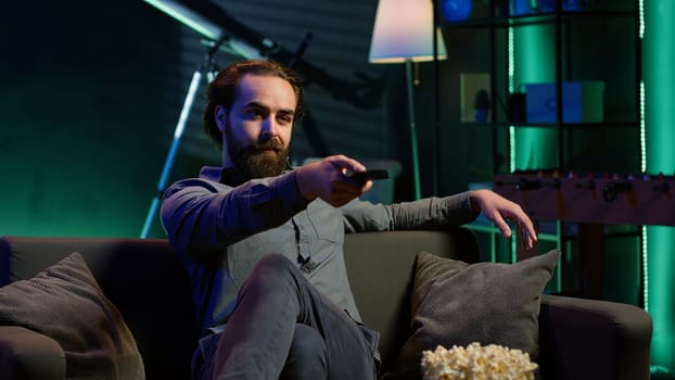 Man taking seat on sofa to watch television, getting entertained in relaxed home environment. Person arriving in apartment sitting down on couch to enjoy movies, turning on TV, eats popcorn, camera A