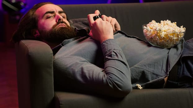 Gamer lounged on couch engaging in multiplayer game using gamepad, eating snack. Person in home theatre playing videogames on gaming console with bowl of popcorn in lap, camera A