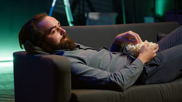 Amused man lounged on couch laughing at funny comedy movie while eating from bowl of popcorn. Cheerful person laying down in apartment amused by hilarious humor in film scene, camera A close up