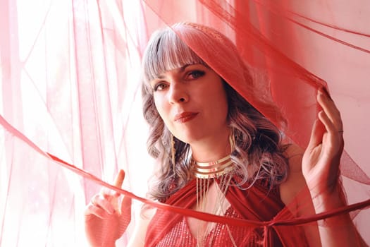 A pretty woman in a red dress looks through a curtain