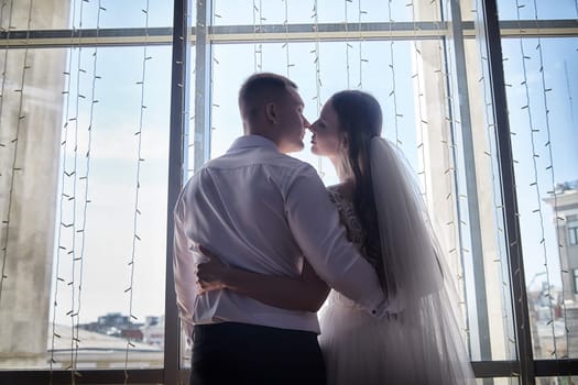 Groom and bride embrace each other, sharing a tender kiss against a bright window adorned with fairy lights, capturing their love.