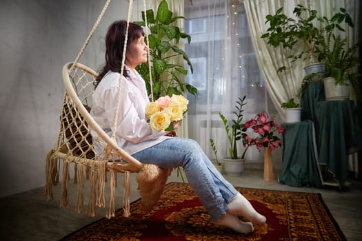 A middle-aged woman poses on a swing inside an apartment, holding roses and smiling softly in a warm, inviting space.