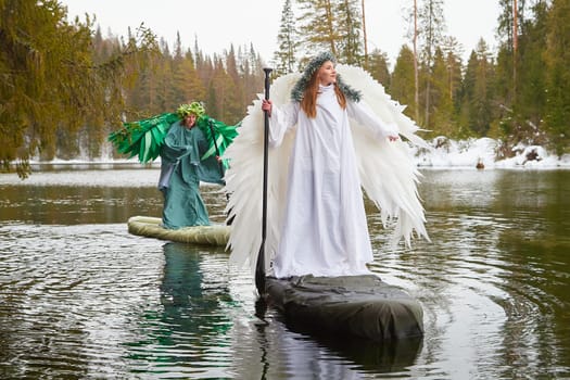 Two girls in white and green dresses gracefully role-play the transition from winter to spring at a tranquil lake surrounded by nature.