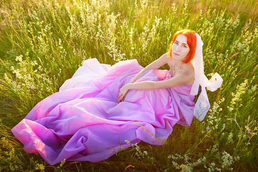 A model with vibrant orange hair sits gracefully on a blanket of pink fabric in a sunlit grassy field. Wildflowers surround her, creating a serene atmosphere at dusk.