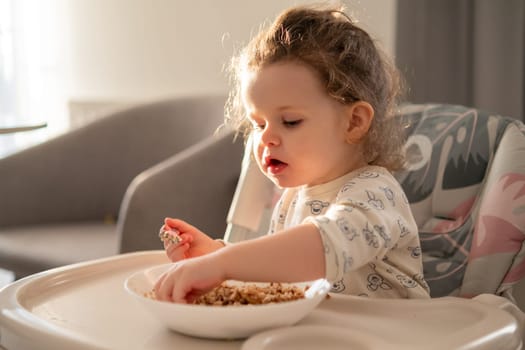Small curly girl eating cereal at the table in a childs high chair. Natural light and warm home atmosphere.