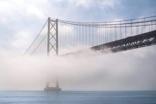 Iconic 25 de Abril suspension bridge, Lisbon, Portugal, in morning fog on sunny day over calm Tagus river waters. Misty atmosphere, peaceful, quiet scene, mist roll in over waters, riverscape, beauty in nature, tourist attraction, travel destination