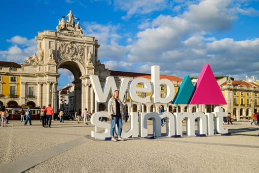 Nov 13, 2024 - Lisbon, Portugal: A female adult posing for a photo in front of Web Summit sign installation at Commerce square and Rua Augusta Arch. People walking, international technology conference, discussions, networking, technology, innovation