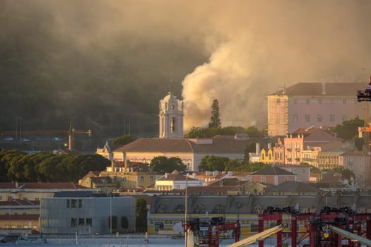 Lisbon church tower in sundown light surrounded by historic buildings and column of rising smoke in air. Soft sunlight, city architecture, beautiful tranquil urban skyline, risk of fires, damage, summer heatwave, emergency, firefighters alert.