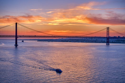 Iconic 25 de Abril bridge in silhouette over Tagus River, Lisbon, Portugal, at sunset with small boat sailing on calm waters. Vibrant orange and purple sky, dramatic backdrop, famous Portuguese landmark, colorful reflections, landscape, summer haze