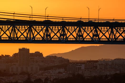 Striking close-up of famous 25 de Abril bridge, its silhouette against bright orange sunset sky. Lisbon, Portugal, cityscape and distant hills in hazy summer evening, blurred vehicles cross bridge, modern buildings, industrial structure, urban life