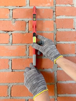 A builder uses a bubble level to check a new red brick wall indoors. Construction and renovation of buildings. Vertical photo with hands and level on the wall.