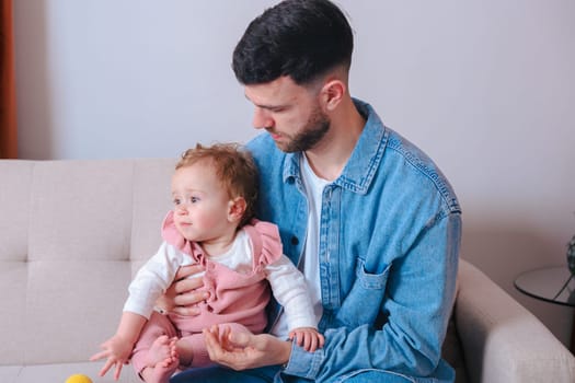 Young father in denim shirt gently holding his baby daughter while sitting on a sofa. The baby looks away curiously, capturing a calm and intimate moment of parenting at home.