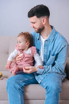 Vertical portrait Young father in denim shirt gently holding his baby daughter while sitting on a sofa. The baby looks away curiously, capturing a calm and intimate moment of parenting at home.