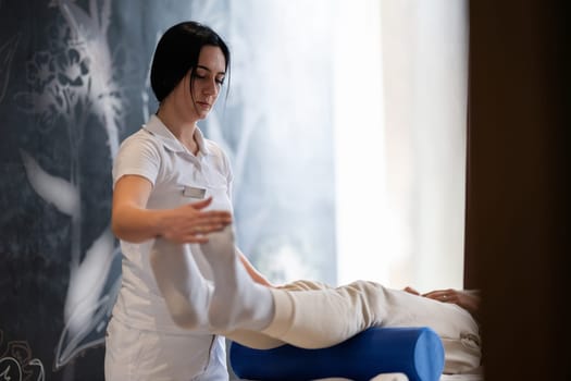 A therapist guiding a patient through leg exercises with the help of a blue foam roller in a therapy clinic.