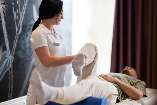 A therapist guiding a patient through leg exercises with the help of a blue foam roller in a therapy clinic.