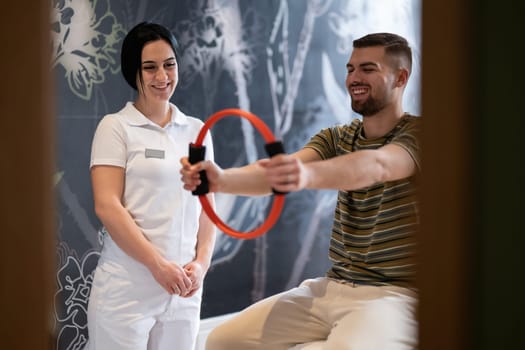 A male patient performing arm exercises using a resistance ring under supervision during a rehabilitation session.