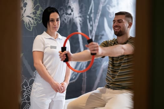 A male patient performing arm exercises using a resistance ring under supervision during a rehabilitation session.