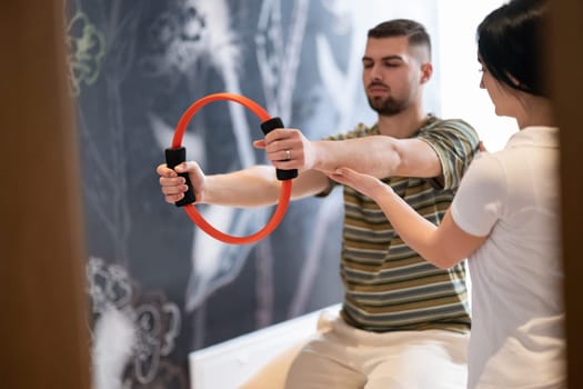 A male patient performing arm exercises using a resistance ring under supervision during a rehabilitation session.