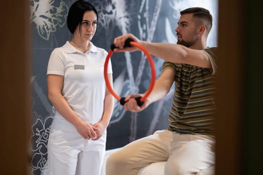 A male patient performing arm exercises using a resistance ring under supervision during a rehabilitation session.