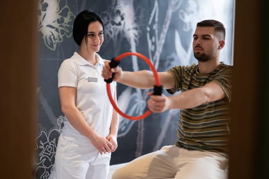 A male patient performing arm exercises using a resistance ring under supervision during a rehabilitation session.