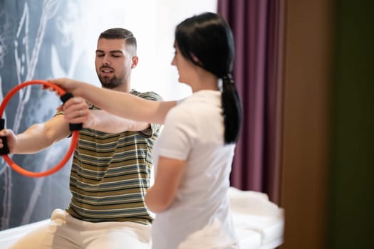 A male patient performing arm exercises using a resistance ring under supervision during a rehabilitation session.