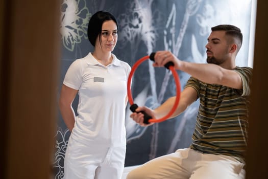 A male patient performing arm exercises using a resistance ring under supervision during a rehabilitation session.