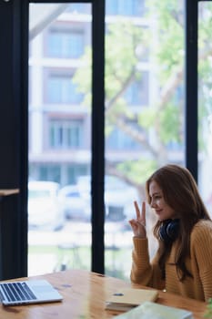Young woman sits at a desk showing the peace sign near her laptop computer..
