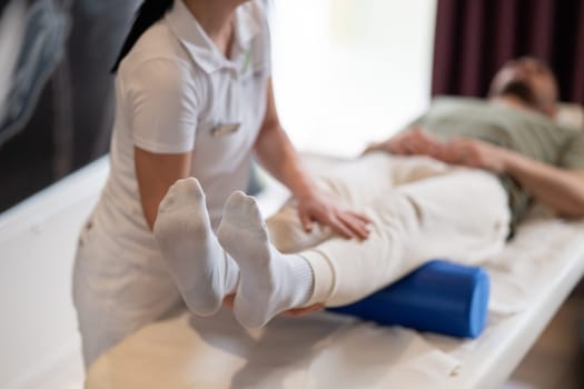 A therapist guiding a patient through leg exercises with the help of a blue foam roller in a therapy clinic.