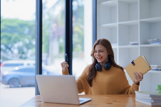 Young woman celebrates success with laptop and book at her bright workspace.