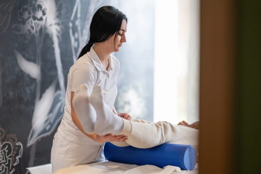 A therapist guiding a patient through leg exercises with the help of a blue foam roller in a therapy clinic.