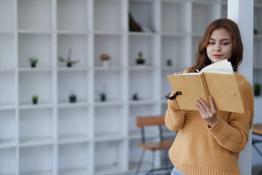 Young woman in sweater reads book near modern bookshelf at cozy home..