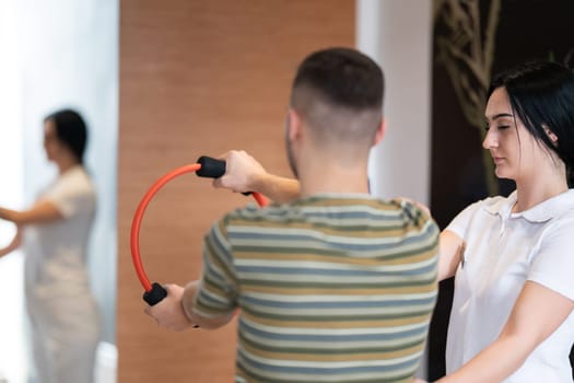A male patient performing arm exercises using a resistance ring under supervision during a rehabilitation session.